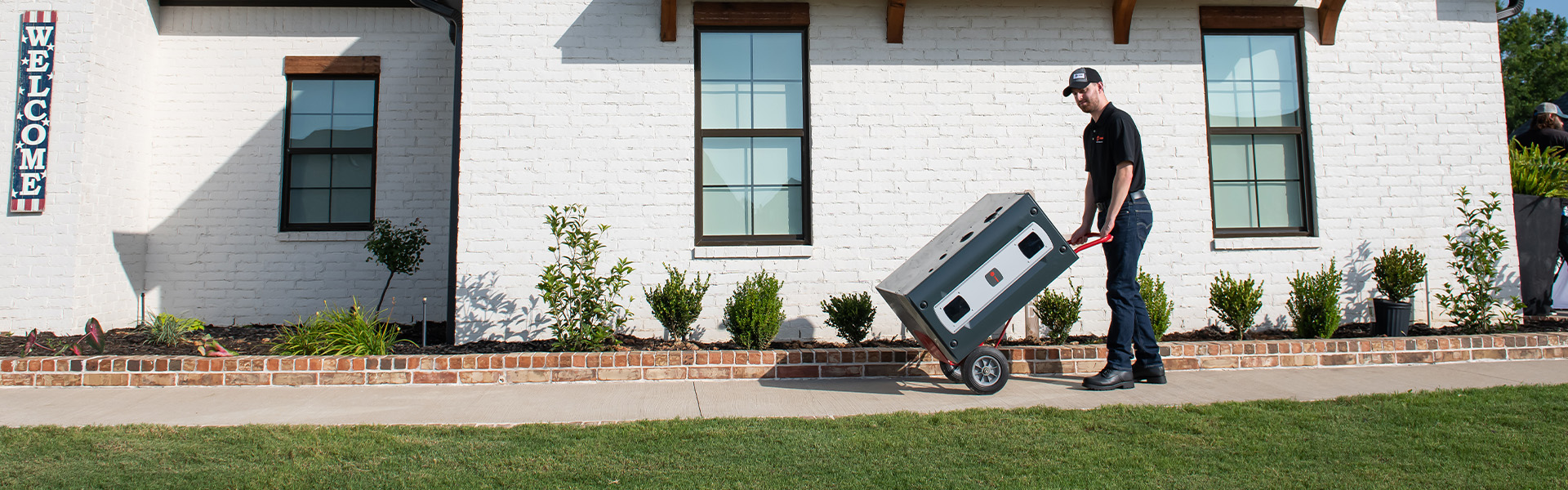 A man pushes a portable generator on a hand truck along a sidewalk in front of a white brick house with landscaping.